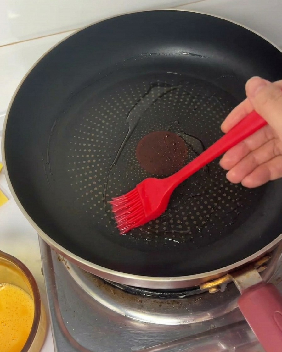 A hand using a red silicone brush to spread cooking oil in a black non-stick frying pan.