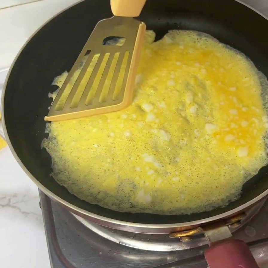 A thin yellow egg omelet being cut into four pieces using a metal spatula in a black frying pan.