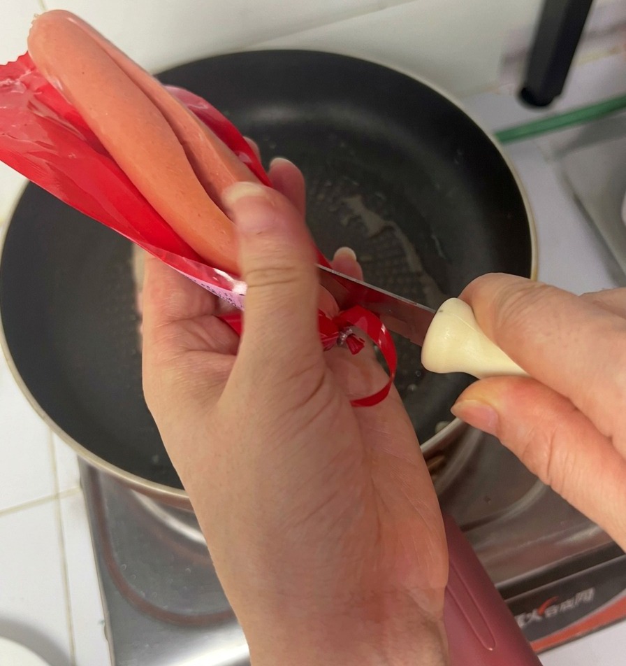 Hands using a knife to open a red ham sausage package over a frying pan containing cooking bacon.