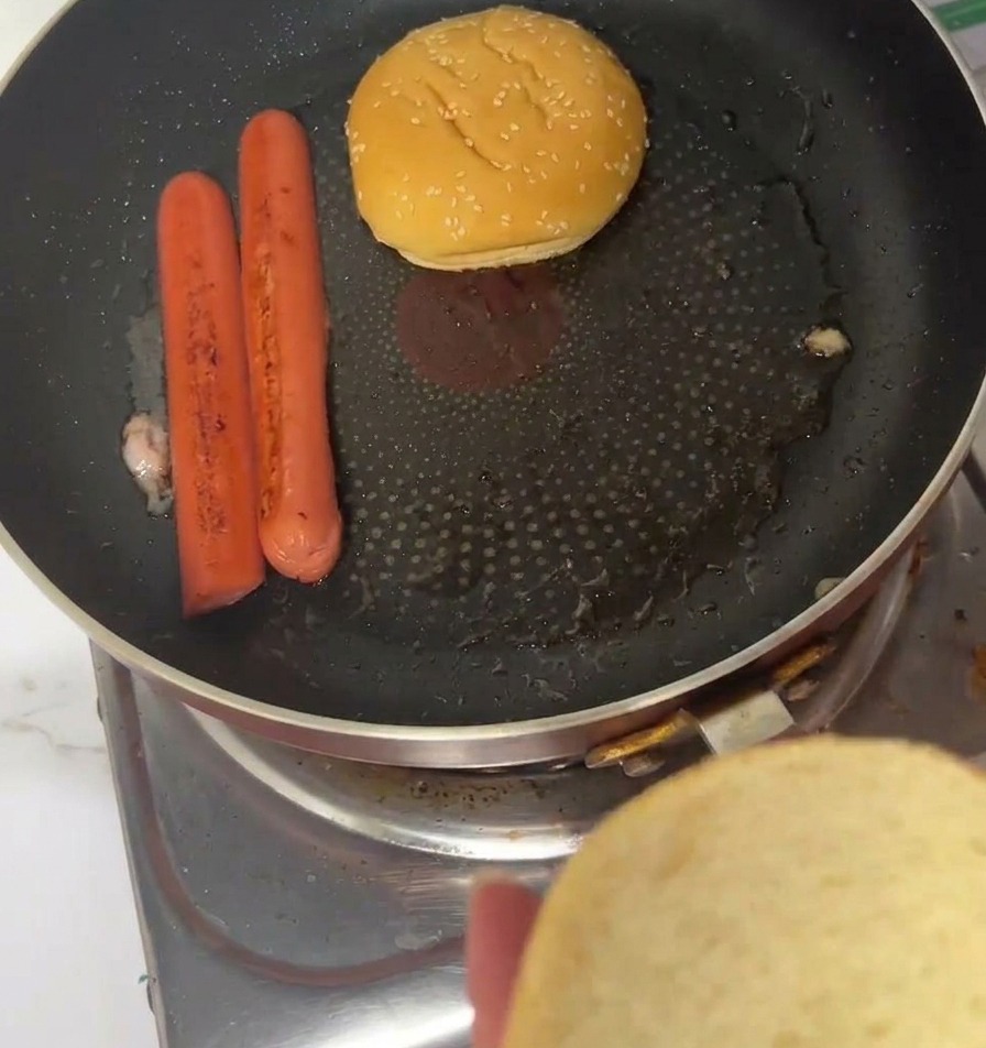 Sesame burger buns being toasted face-down in a frying pan next to cooked whole sausages.