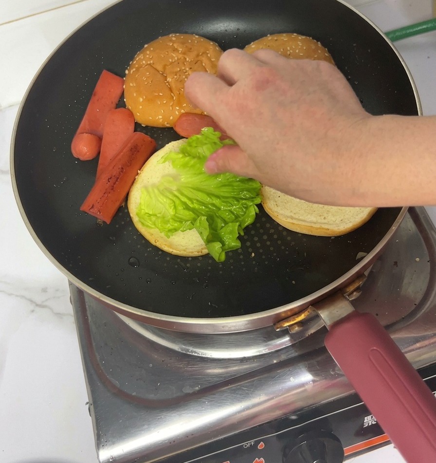 A hand placing a fresh green lettuce leaf onto a toasted bun inside a frying pan.