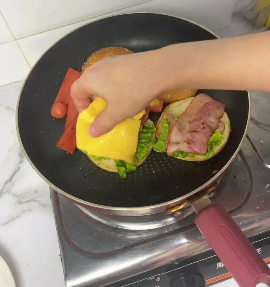 A hand placing a square slice of yellow cheese onto a partially assembled burger with lettuce and bacon inside a frying pan.