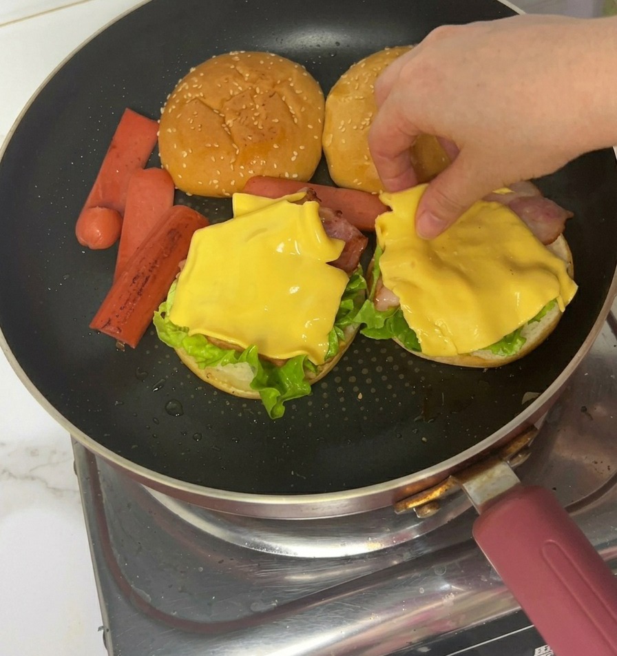 Hands using tongs to place cooked ham slices over the cheese layer on a partially assembled burger.