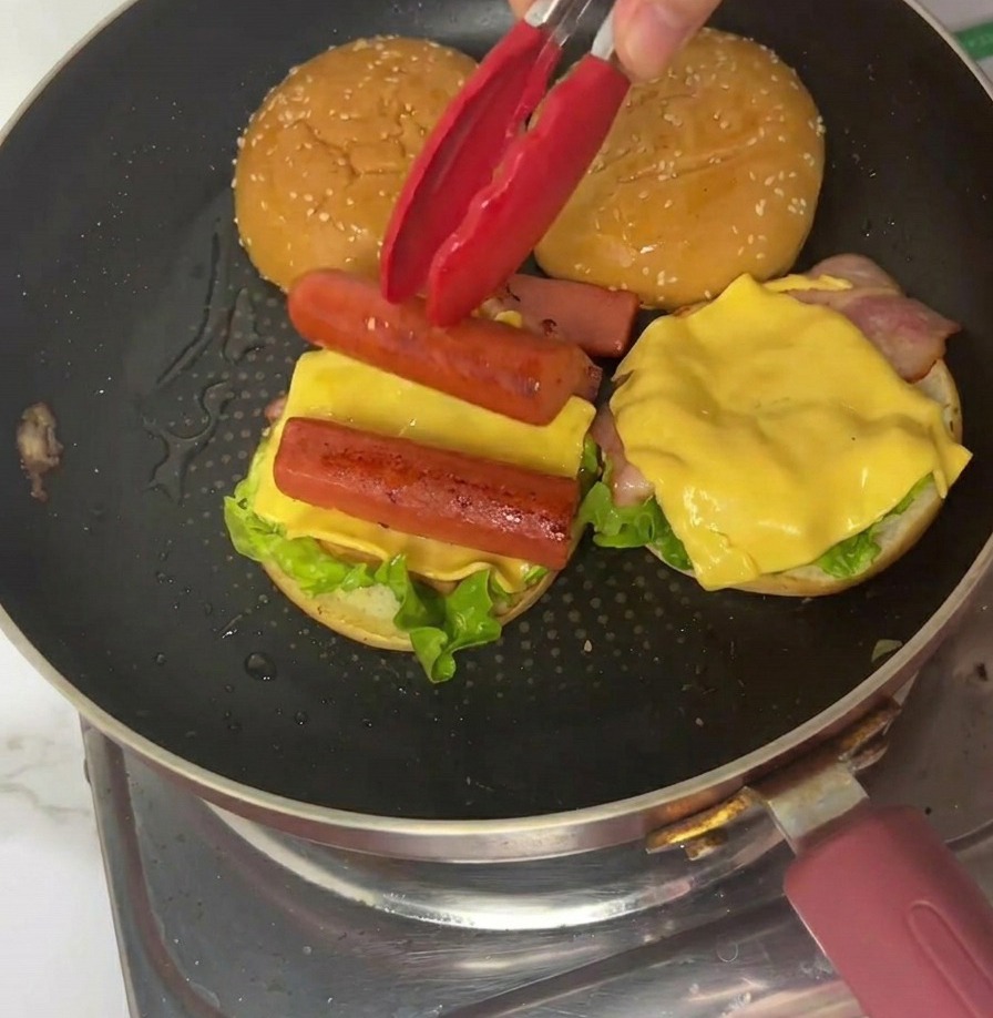 A quarter of a thin egg omelet being placed onto a burger assembly using red tongs in a frying pan.