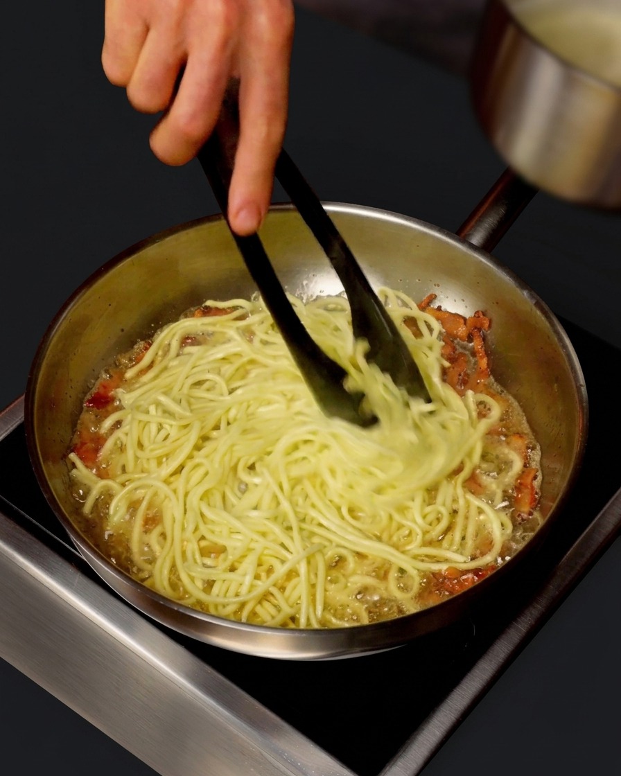 A small pot pouring hot pasta water into a skillet of spaghetti and guanciale while being tossed with tongs.