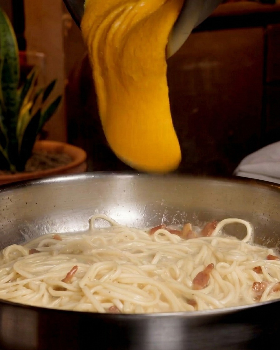 A thick, bright orange egg and cheese mixture being poured from a metal bowl over a pan of spaghetti and guanciale.