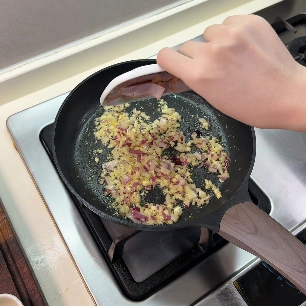 Sliced bacon being slid from a small plate into a frying pan containing sautéed onions and garlic.