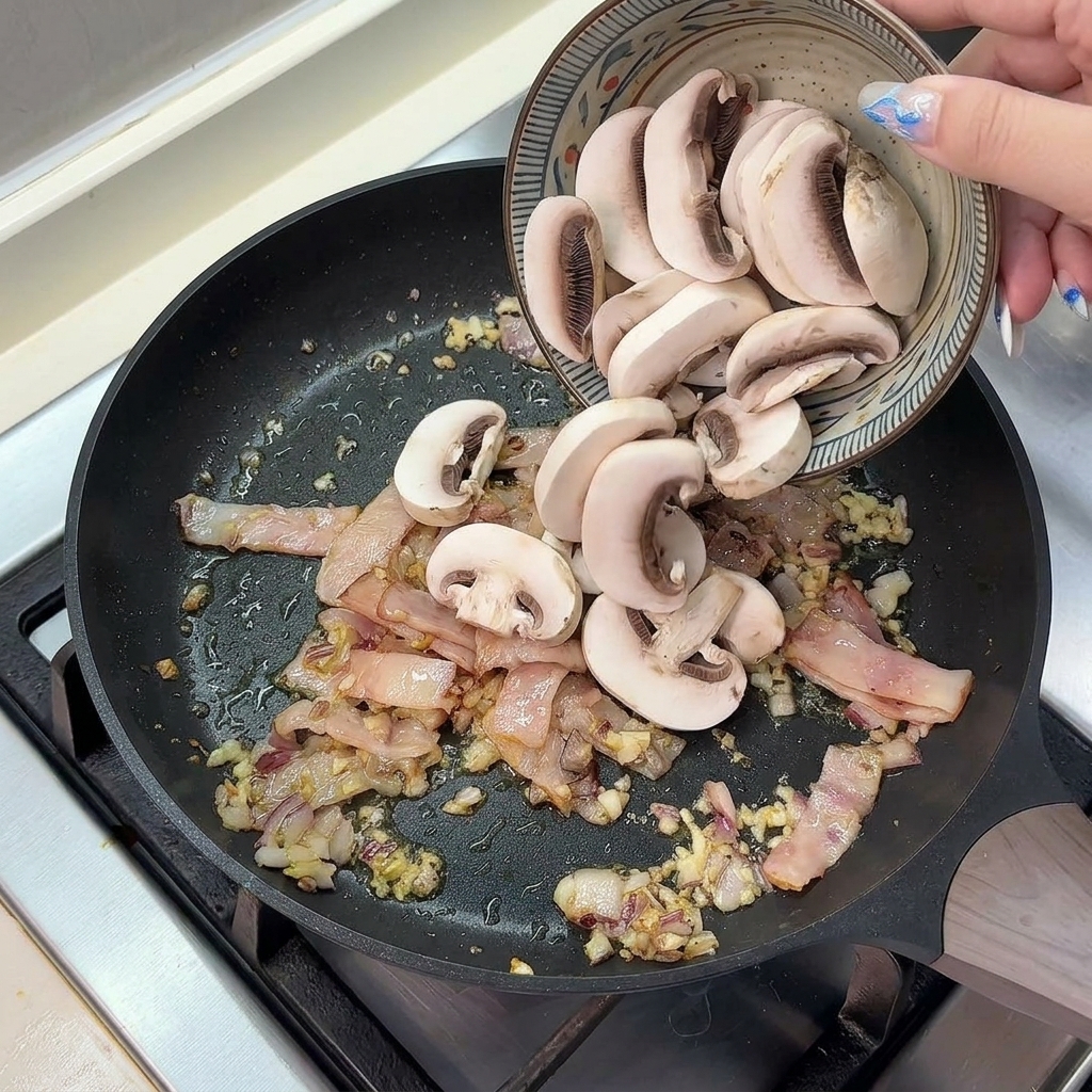 Freshly sliced button mushrooms being poured from a ceramic bowl into a hot pan containing cooked bacon, onions, and garlic.