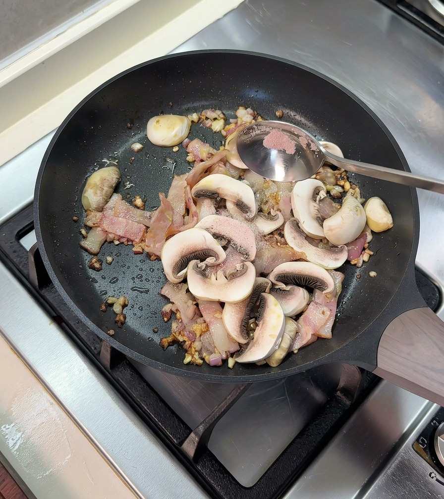 Adding a spoonful of pink salt to a frying pan with cooked bacon and sliced button mushrooms.