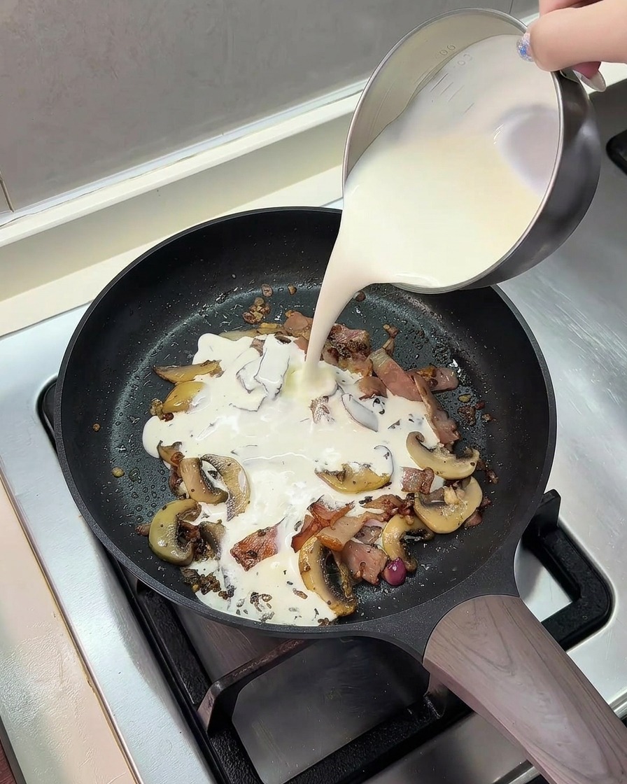 Pouring a mixture of milk and heavy cream from a metal pitcher into a frying pan with cooked bacon and mushrooms.