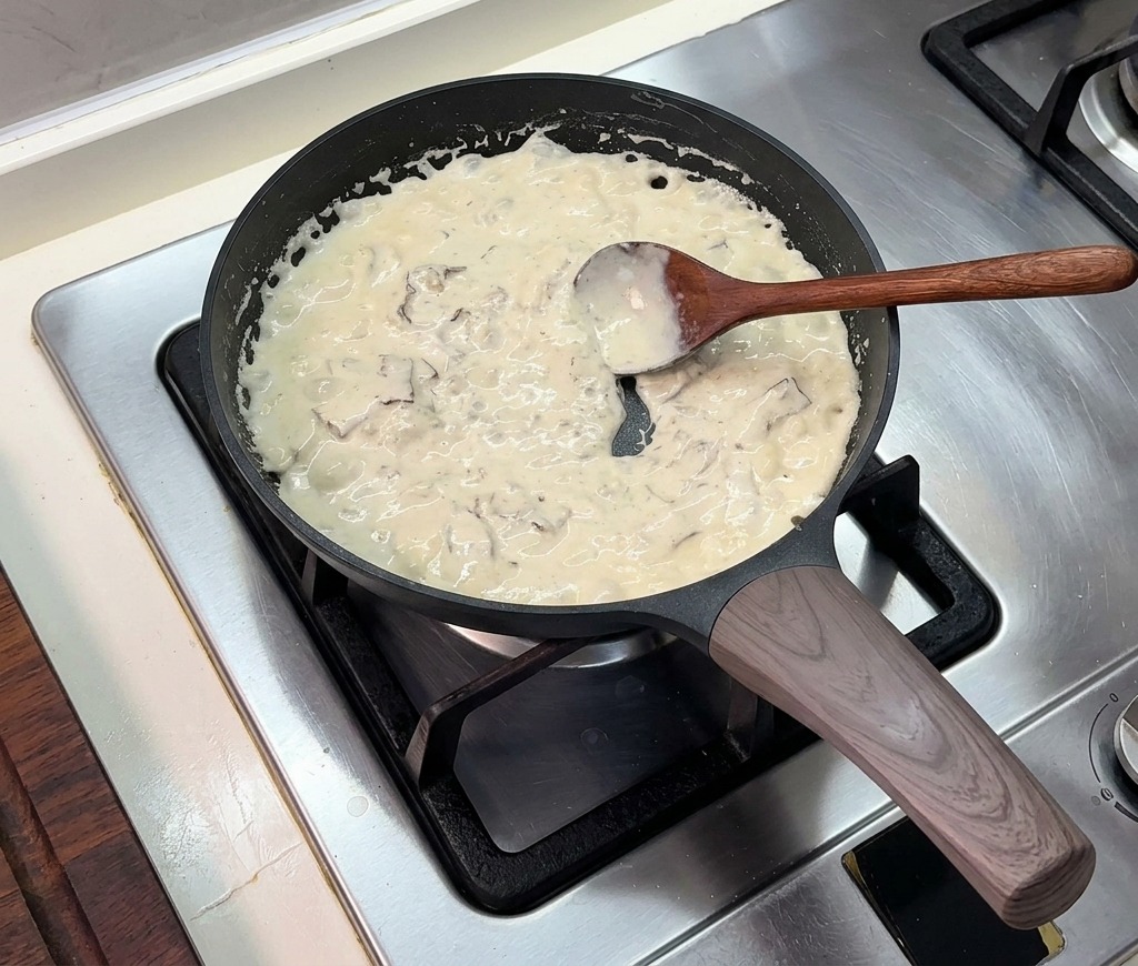 A thick, bubbling cream sauce simmering in a black frying pan, being stirred with a wooden spoon.