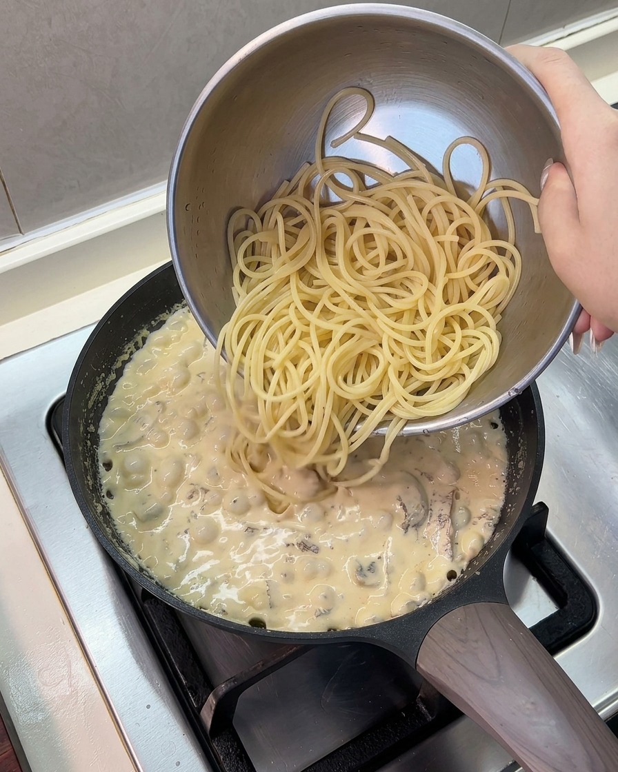 Cooked spaghetti noodles being poured from a stainless steel bowl into a pan of simmering cream sauce.