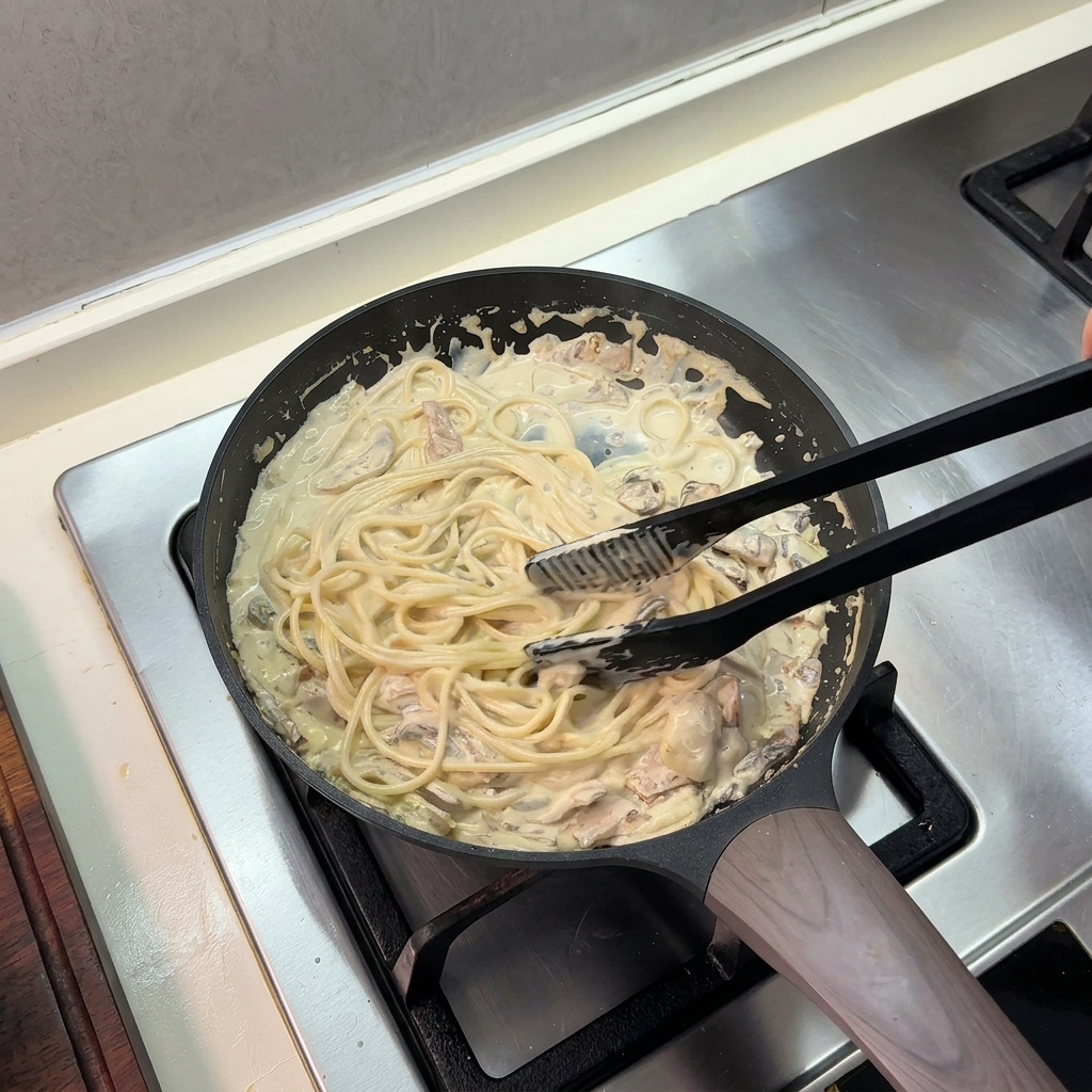 Black kitchen tongs tossing cooked spaghetti in a rich cream sauce inside a frying pan.