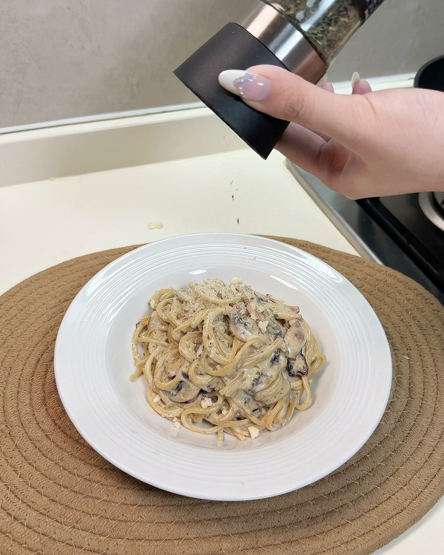 A hand using a grinder to sprinkle cheese and herbs over a warm plate of creamy mushroom and bacon spaghetti.