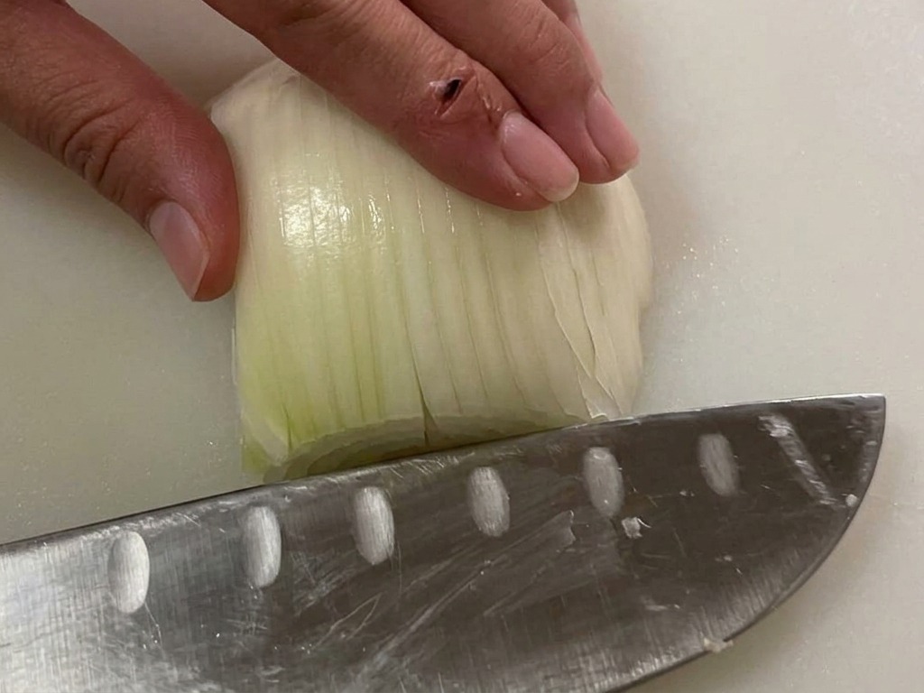 Hand holding a white onion half on a cutting board while a knife prepares to dice it.