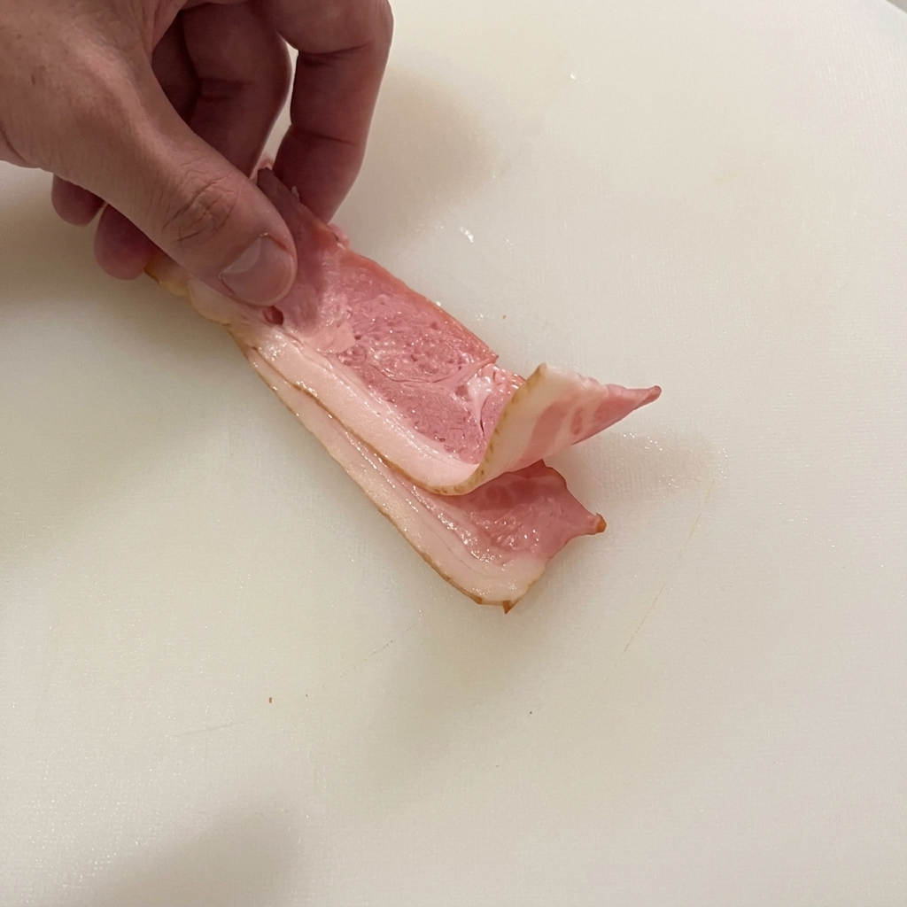 Hand folding two strips of raw bacon on a white cutting board, preparing to chop them.