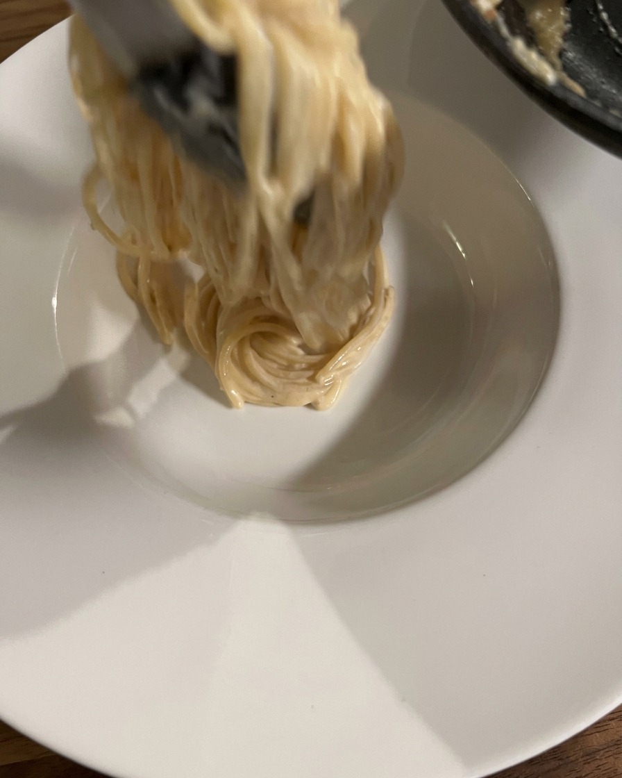 Tongs lifting and twirling a portion of creamy spaghetti into a clean white ceramic serving bowl.