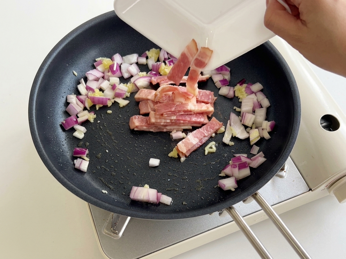 Sliced bacon being poured from a white plate into a frying pan with diced red onions and garlic.