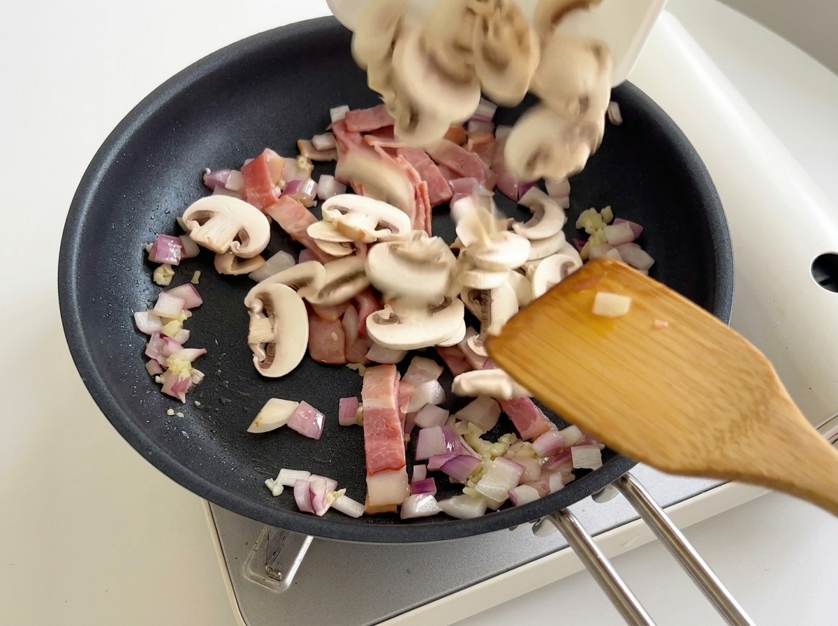 Sliced button mushrooms being added from a plate into a frying pan containing bacon and onions.