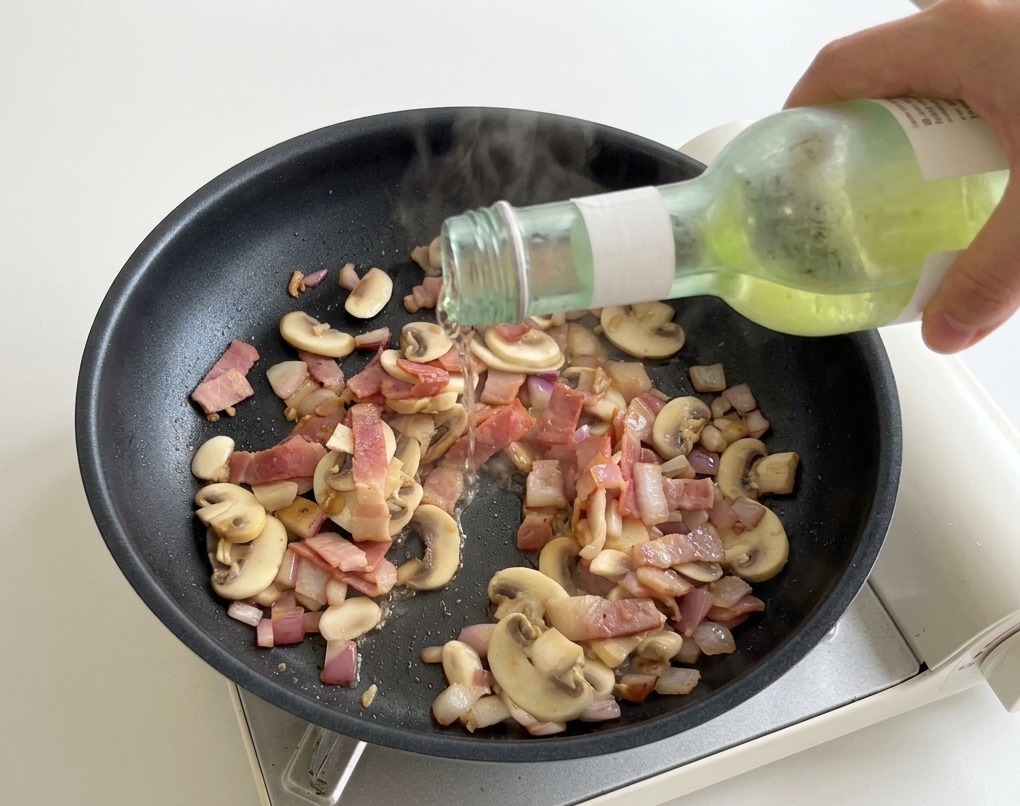 A hand pouring white wine from a small green bottle into a hot skillet with sliced mushrooms and bacon pieces.