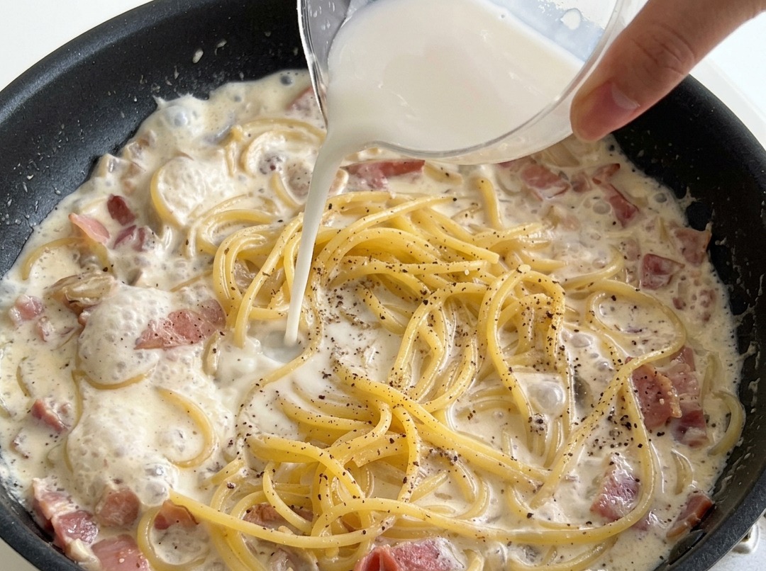 Pouring a white water starch mixture from a small measuring cup over cooked spaghetti in a pan with creamy bacon and mushroom sauce.