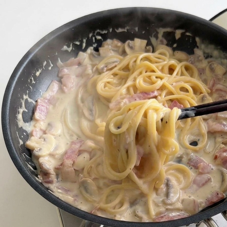 Black chopsticks twirling a portion of creamy spaghetti within a frying pan filled with a rich, bubbling milk sauce, bacon, and mushrooms.