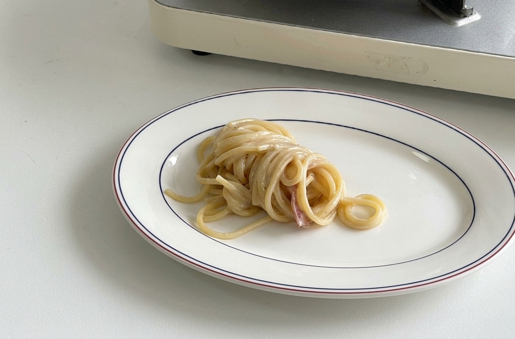 A neatly twirled mound of creamy spaghetti resting in the center of an oval white plate with a dark blue and red rim.