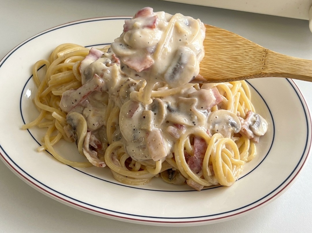 A wooden spoon lifting a hearty scoop of creamy sauce with bacon and mushrooms, pouring it over a serving of spaghetti on a white plate.