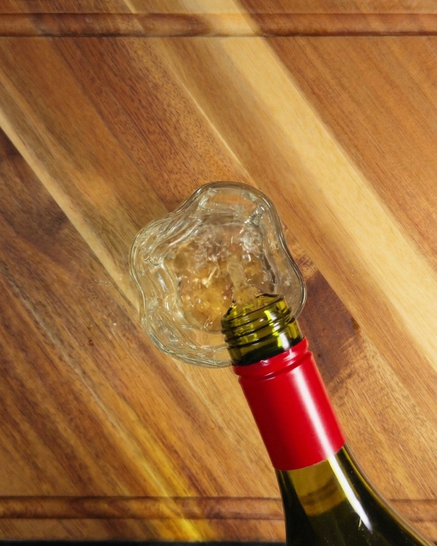 White wine being poured from a green glass bottle into a small clear glass on a wooden surface.