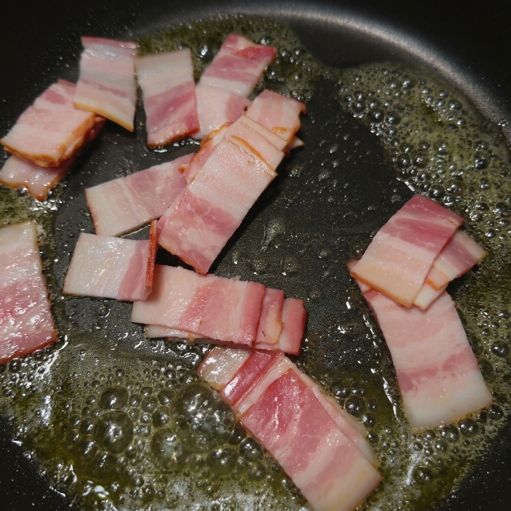 Small strips of bacon sizzling and browning in a black frying pan filled with hot rendered fat.