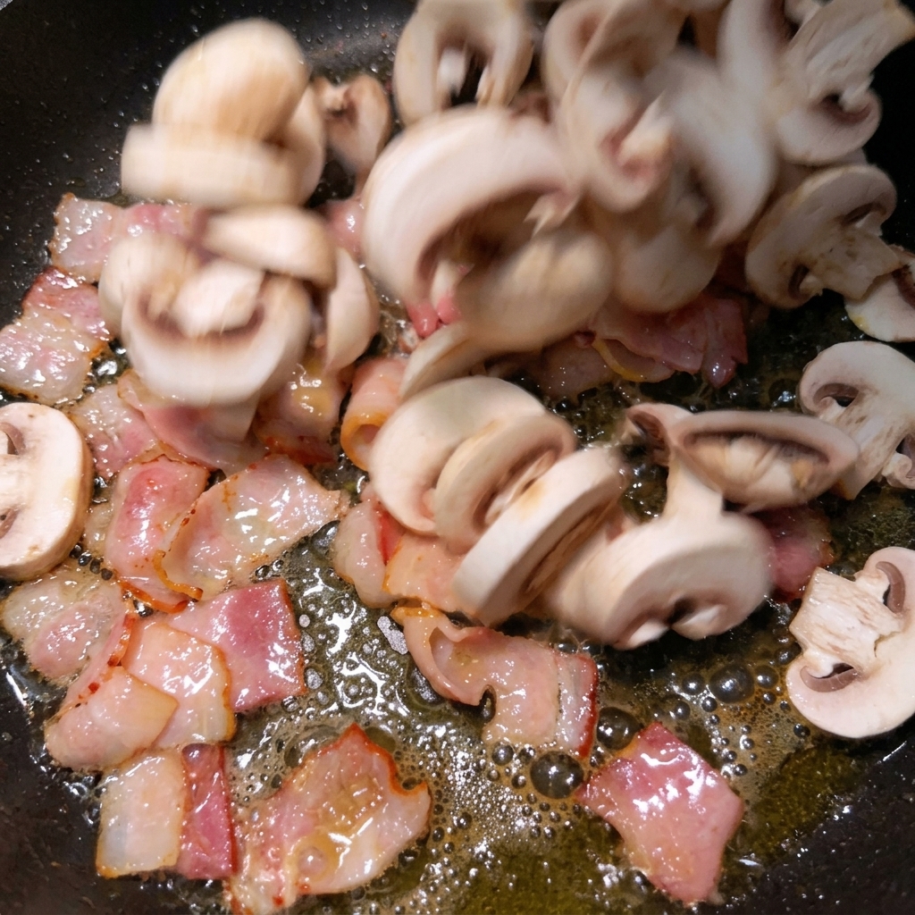 Sliced white mushrooms falling into a hot pan filled with sizzling, rendered bacon pieces.
