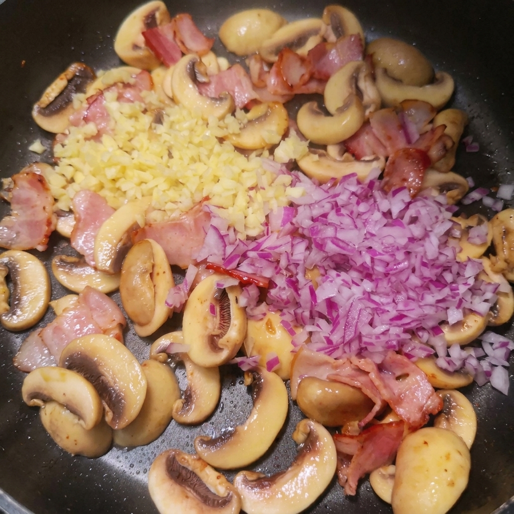 Freshly minced red onion and garlic piled on top of cooked bacon and sliced white mushrooms in a pan.