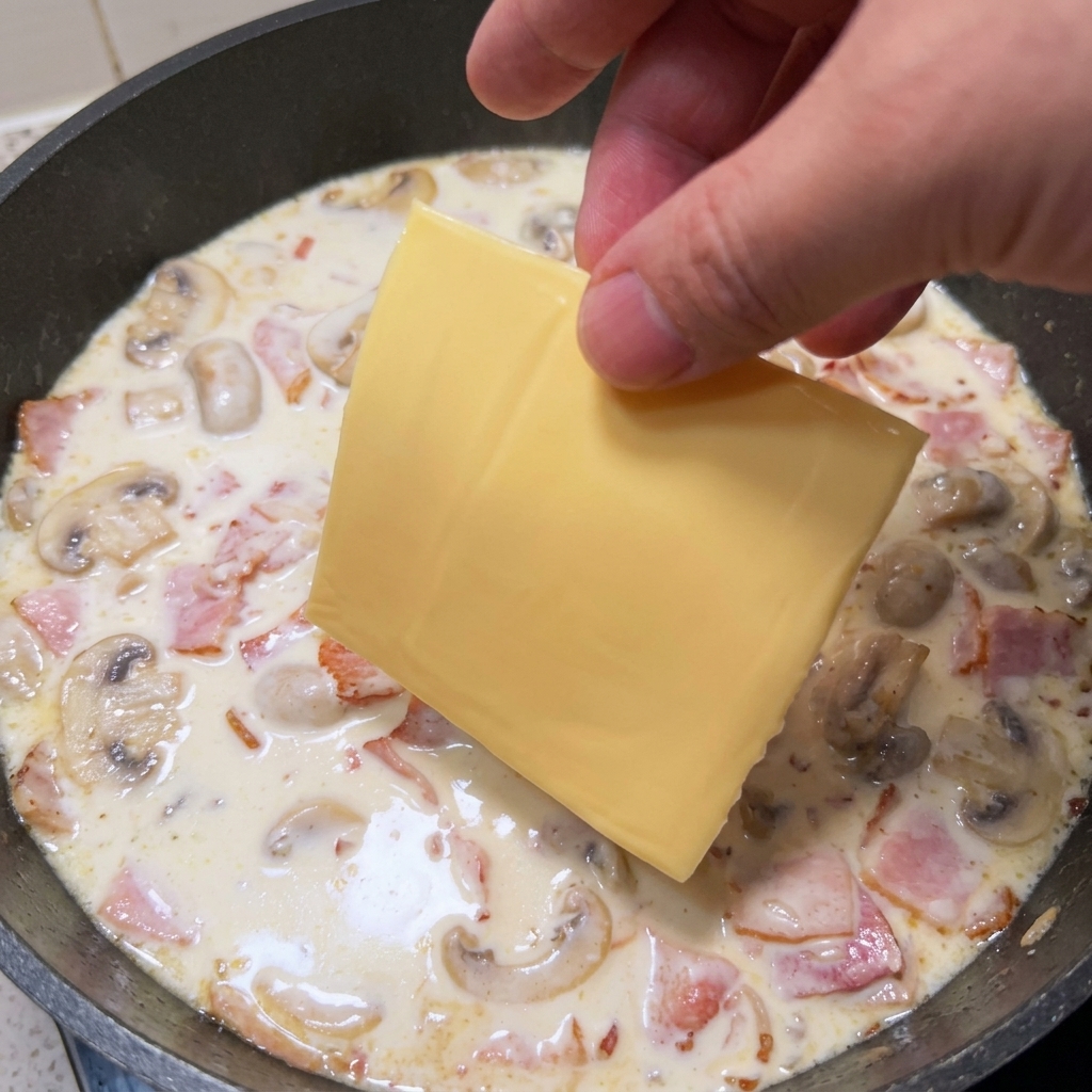 A hand placing a square slice of yellow cheese into a hot pan filled with creamy white sauce, sliced mushrooms, and bacon bits.