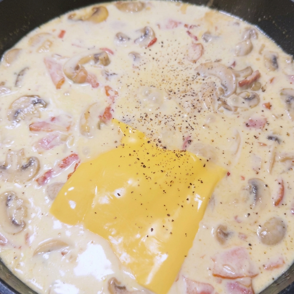 A slice of yellow cheese melting in a pan of creamy mushroom and bacon sauce, generously seasoned with freshly ground black pepper.
