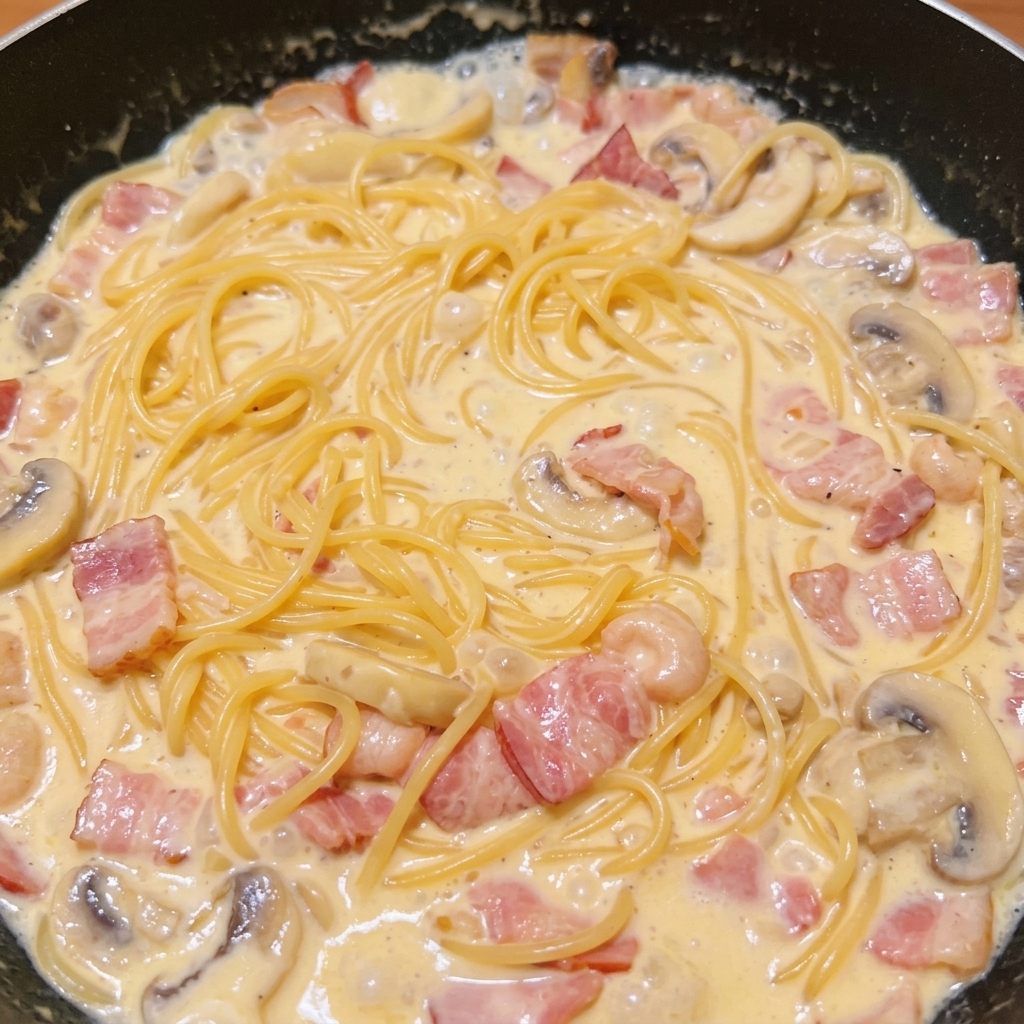 Cooked spaghetti noodles thoroughly mixed into a rich, creamy sauce with rendered bacon bits and sliced mushrooms in a dark skillet.