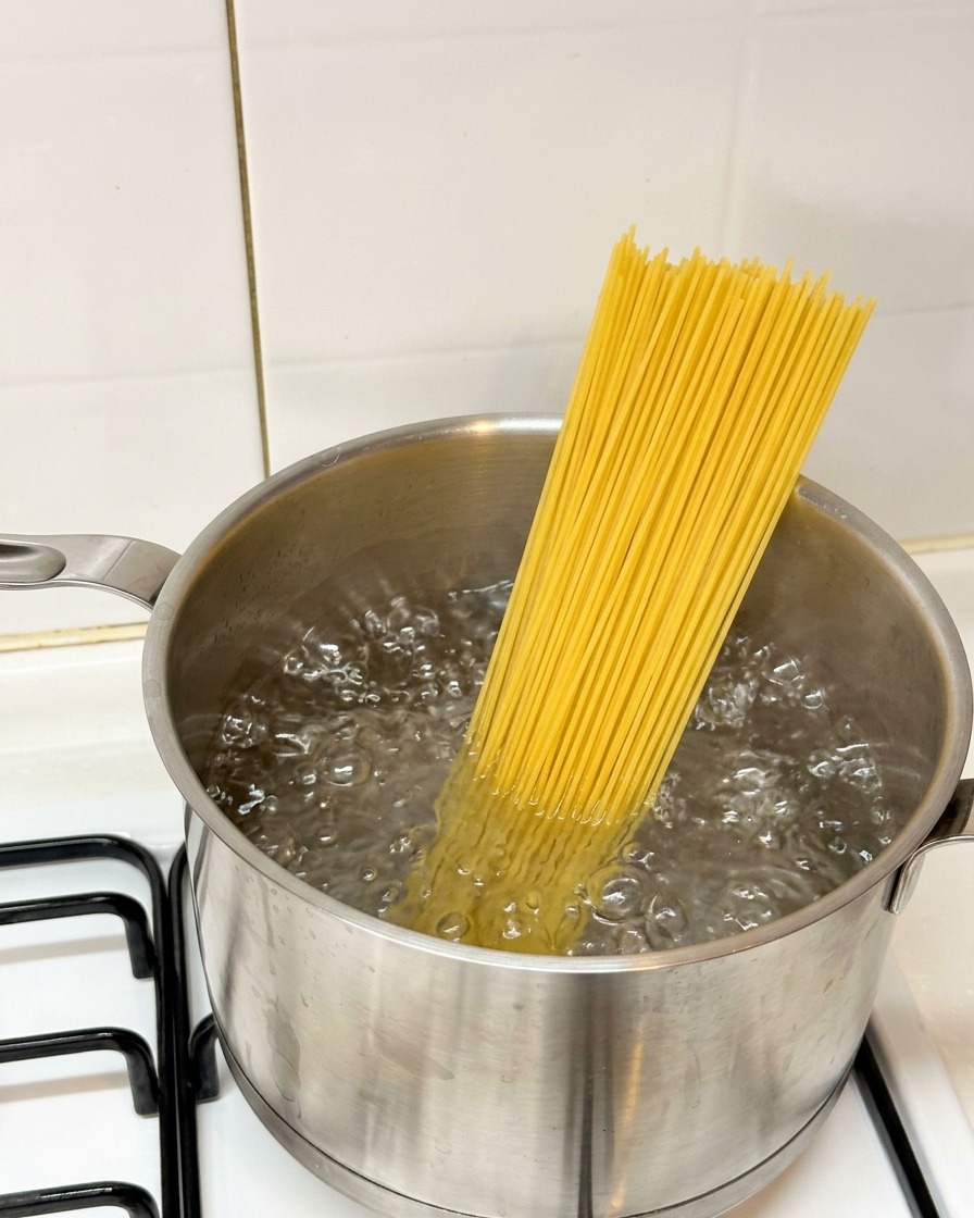 Dry spaghetti pasta being placed into a pot of vigorously boiling water on a stovetop.