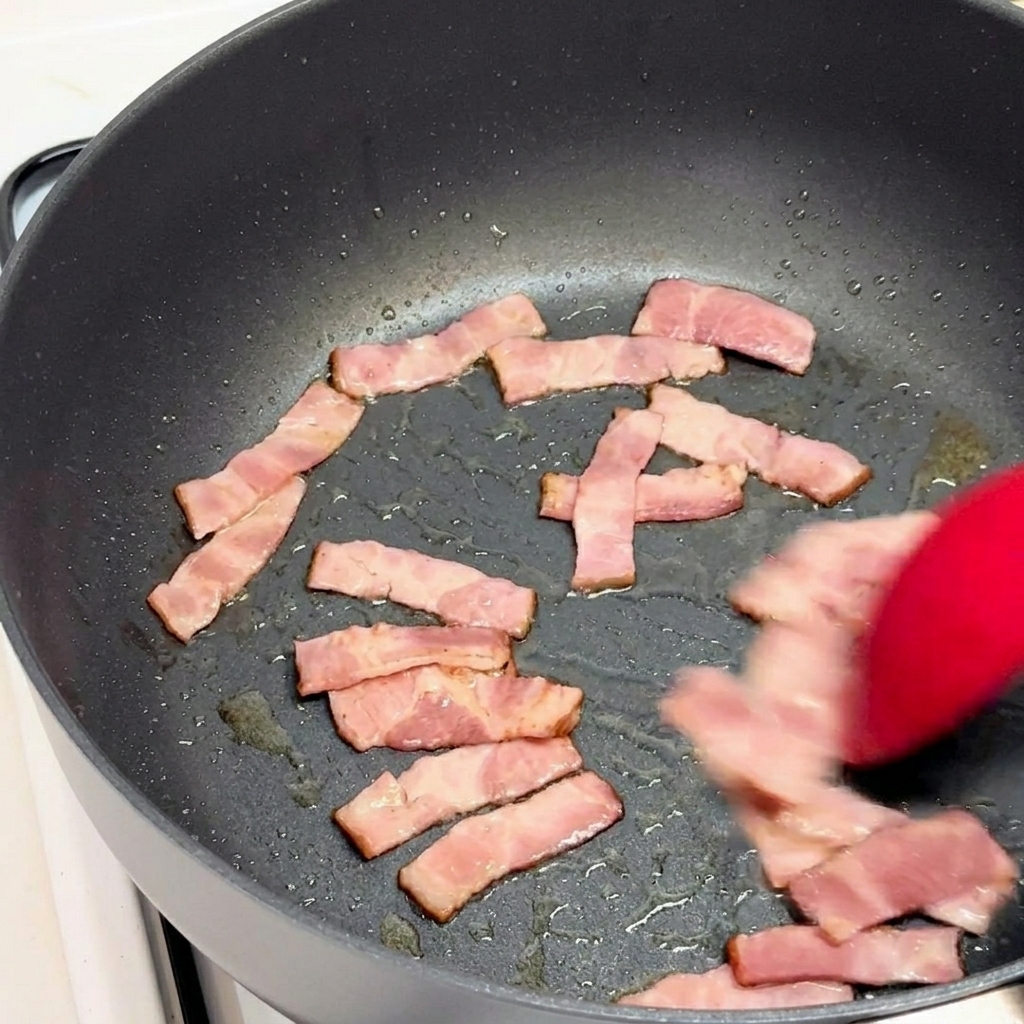 Pieces of bacon being stir-fried with a red silicone spatula in a black skillet.