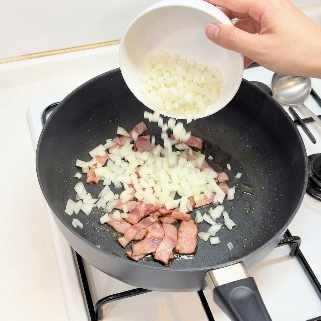 A hand pouring diced white onions from a white bowl into a pan containing cooked bacon pieces.