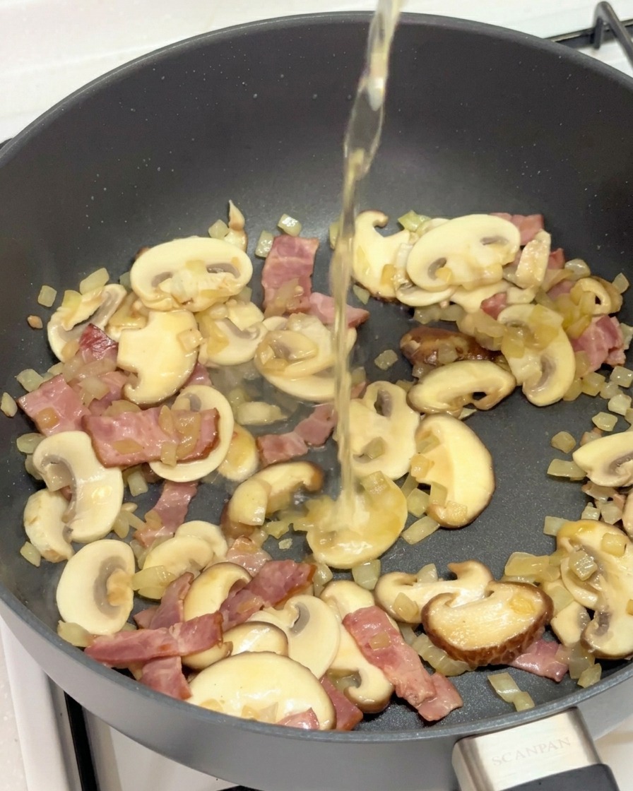 A clear liquid being poured into a pan filled with sautéed mushrooms, bacon, and onions.