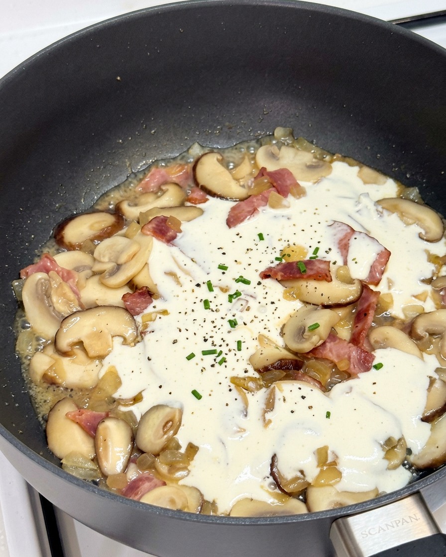 Heavy cream simmering in a skillet with mushrooms and bacon, sprinkled with black pepper and fresh herbs.
