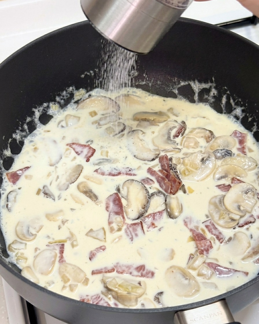 A person using a grinder to crack fresh black pepper into a skillet filled with a creamy mushroom and bacon sauce.