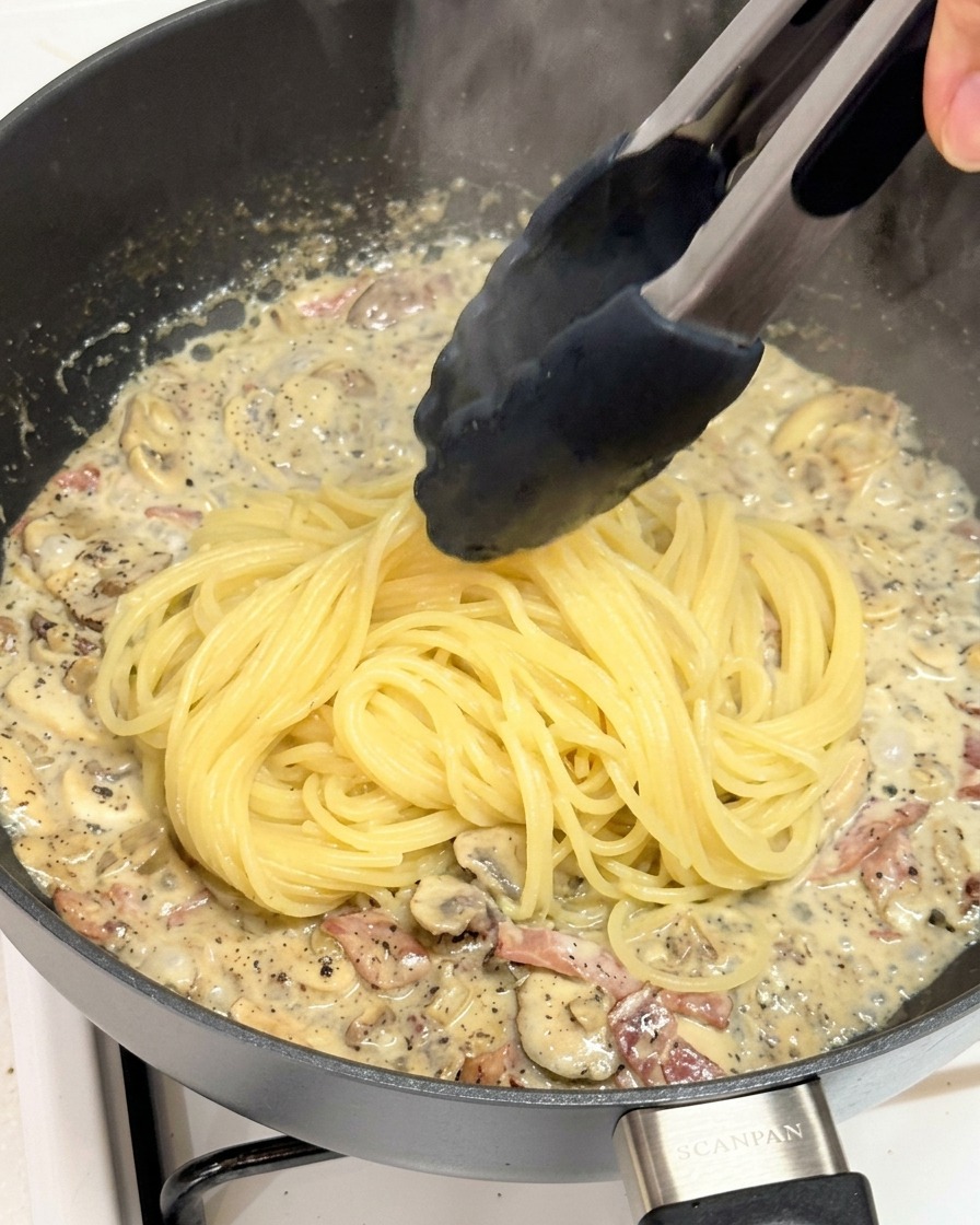 Tongs dropping freshly boiled spaghetti noodles into a skillet filled with a creamy, rich truffle mushroom sauce.