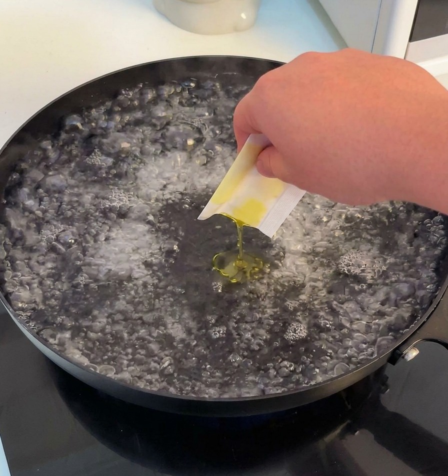 A hand pouring salt and olive oil from a small packet into a wide pan of vigorously boiling water.