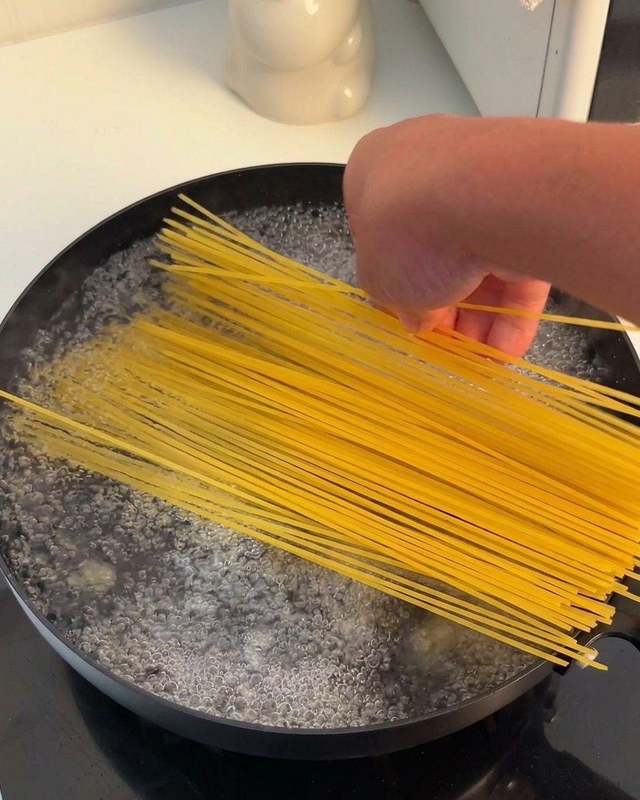 A hand placing a bundle of dry yellow spaghetti noodles into a black pan filled with actively boiling water.