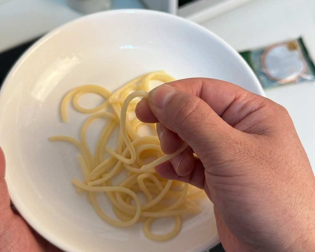 A hand pinching a strand of cooked spaghetti over a white bowl to check if it is tender.