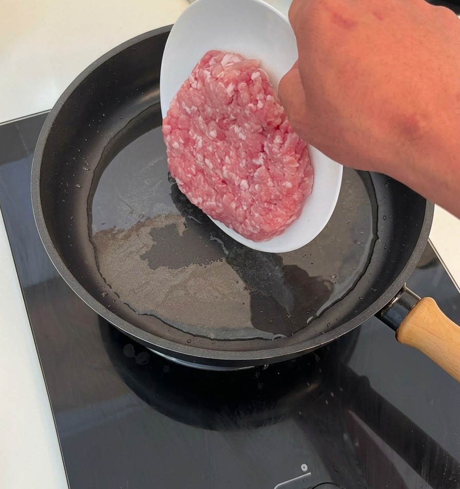 A hand sliding a portion of raw minced meat from a white plate into a hot frying pan coated with cooking oil.