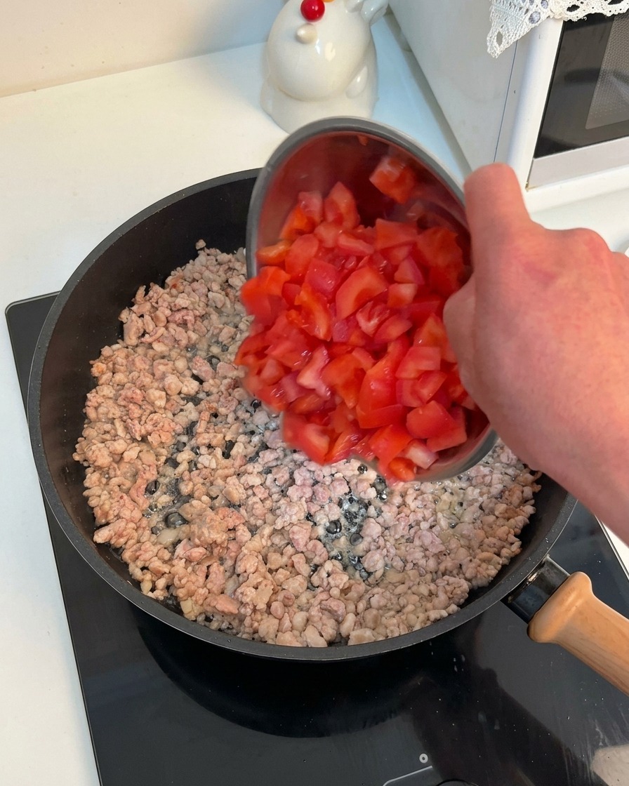 A hand pouring a bowl of bright red diced tomatoes into a hot pan containing browned minced meat.
