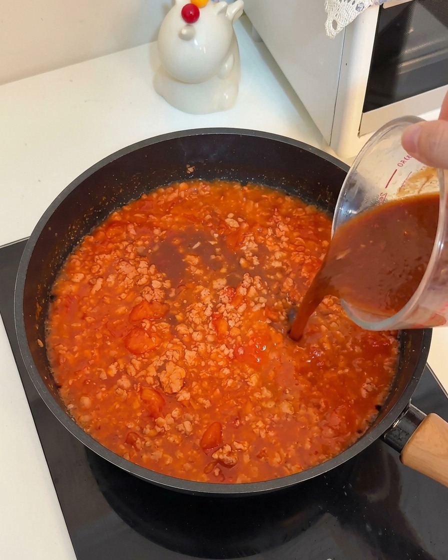 A dark liquid sauce being poured from a glass measuring cup into a pan of simmering meat and tomatoes.
