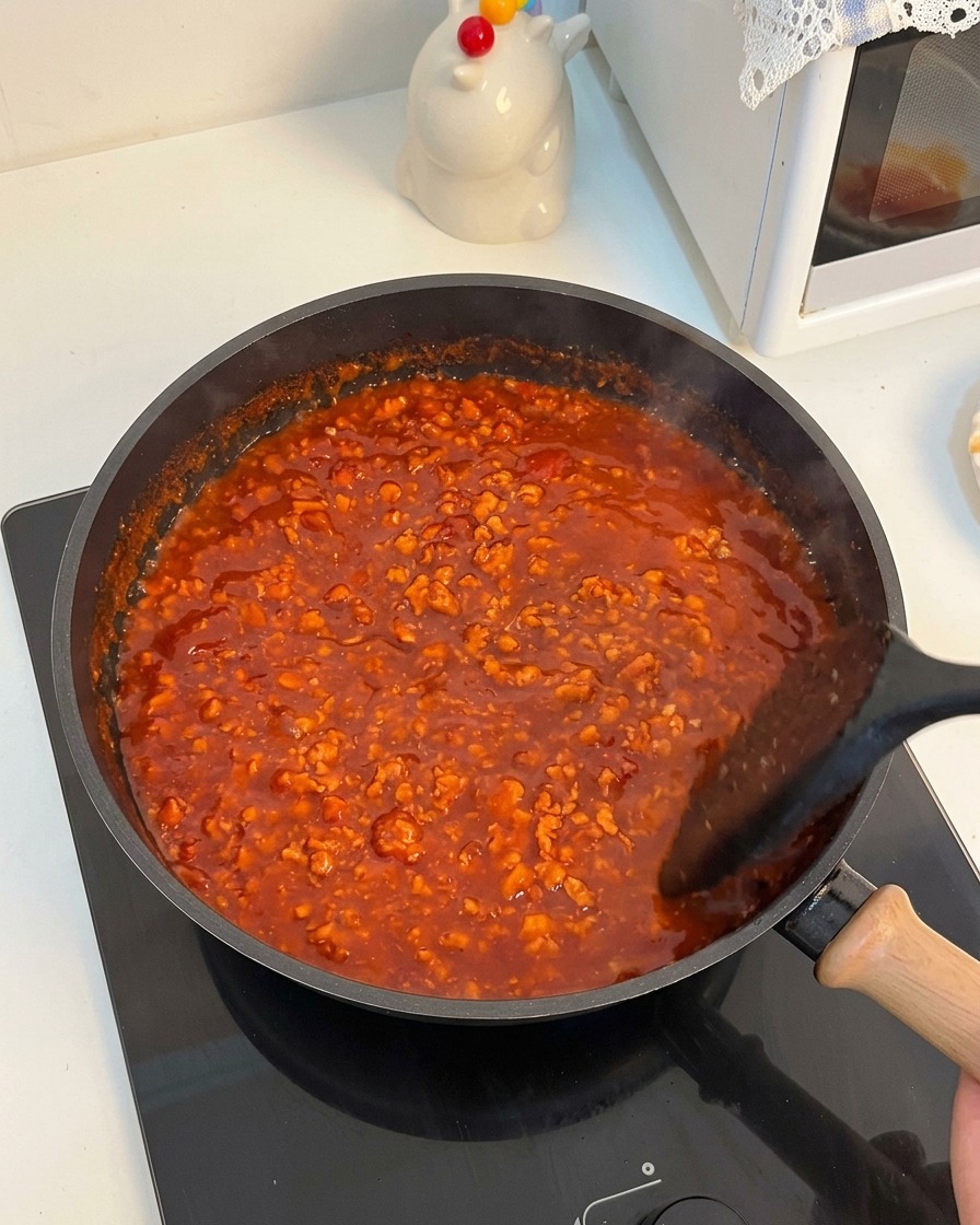 Rich, vibrant red tomato meat sauce bubbling vigorously in a frying pan while being stirred with a black spatula.