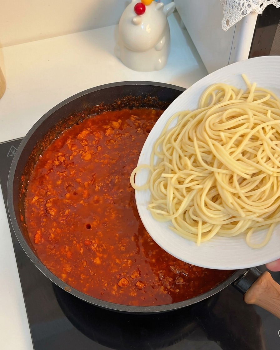 Cooked spaghetti noodles being gently slid from a white plate into a pan filled with rich tomato meat sauce.